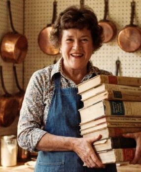 Julia Child holding a stack of her cookbooks in her kitchen