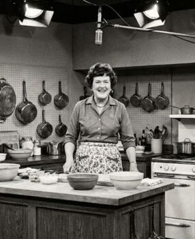 Julia Child standing in her TV kitchen set smiling at camera in the 1960s