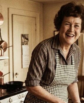 Julia Child smiling warmly in her 1960s kitchen with copper pots on pegboard behind her